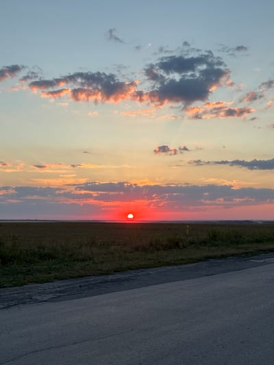The image depicts a rural landscape at sunset. The sun, a vibrant red sphere, is low on the horizon, casting a warm glow across the sky. Streaks of pink and orange clouds are visible, interspersed with darker, shadowed clouds. The foreground features a dark asphalt road, with a grassy verge and a field of dry vegetation stretching towards the horizon. In the distance, the land appears flat, with a faint suggestion of buildings or structures. A yellow marker is visible in the field on the right side of the image. The scene is devoid of people or any signs of recent activity. The overall impression is one of tranquility and natural beauty.