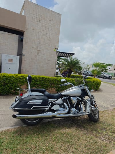 A silver and black motorcycle is parked on a grassy area next to a paved walkway. The motorcycle has saddlebags and a backrest. Behind the motorcycle is a meticulously trimmed green hedge. Beyond the hedge, a building with a light-colored stone facade stands, with a beige overhang on the left side. A white utility box with text is mounted on the wall of the building. Further in the background, palm trees and other vegetation are visible. Across a street, two cars are parked in front of residential buildings. The sky is partly cloudy, suggesting it is daytime. The scene appears to be in a residential or commercial area in Cancún, Mexico. There is no visible activity taking place. The text on the utility box reads: "GRUPO VERMAYA SA DE CV RN. 19-56333. LOTE 13 SP:2192020".