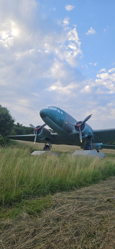 A vintage, twin-engine propeller airplane is parked on a concrete platform amidst tall, dry grass. The aircraft is painted in a camouflage pattern of dark green and blue-grey. Its propellers are visible, with yellow tips. In the background, a dense line of trees forms a natural boundary. The sky above is a light blue, scattered with fluffy white clouds, some of which are illuminated by sunlight. The scene is located in the vicinity of Vyšný Komárnik, Slovakia, a place known for its historical significance related to World War II.