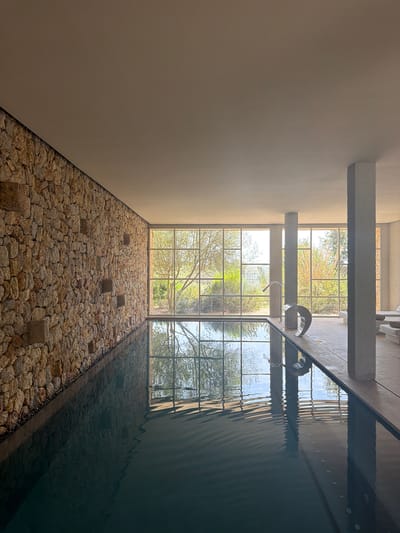 The image depicts an indoor swimming pool area in Artà, Spain. The pool itself is dark blue and partially filled, with ripples on its surface reflecting the light and architecture. To the left, a wall is constructed from natural, irregular stones of varying sizes and colors, creating a rustic texture. Several square, lighter-colored stone niches are embedded in this wall.

Dominating the background is a large expanse of windows, divided into a grid-like pattern by metal frames, offering a view of lush greenery and trees outside. The sunlight streams through these windows, casting geometric shadows on the water's surface and the pool deck.

In the foreground, near the edge of the pool, are two modern, metallic water features: one resembling a sleek spout and the other a curved, sculptural element. Two thick, rectangular concrete pillars stand at intervals, supporting the ceiling. The ceiling is smooth and light-colored, contrasting with the textured walls. To the right of the pool, a section of the floor is visible, with two minimalist lounge chairs placed further back, suggesting a relaxation area. The overall ambiance is serene and contemporary, blending natural materials with modern design.