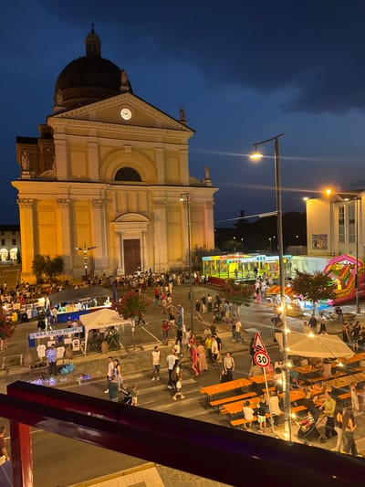 The image depicts an evening outdoor event in the city of Calcio, Italy, centered around the town's church. The church, a prominent yellow building with a large dome and a clock face, is illuminated. The scene is busy with people of all ages, many of whom are standing, walking, and sitting at tables.
In the foreground, a red railing suggests the viewpoint is from an elevated position, possibly a balcony or higher floor. The ground level features market stalls, one with "U.S.O. CALCIO VOLLEY" printed on it, selling merchandise. Further into the scene, there are inflatable attractions, including a bouncy castle, and a colorful trampoline-like structure labeled "TAPPETI ELASTICI". Orange tables and benches are set up, indicating a dining or gathering area, with many people seated and interacting.
Several streetlights and lights from the stalls illuminate the area, creating a lively evening atmosphere. A "30" speed limit sign is visible, suggesting the event is taking place in a public square or street that is temporarily closed or managed for the occasion. The sky is a deep blue, indicating it is dusk or early night. The overall impression is of a community festival or fair.
