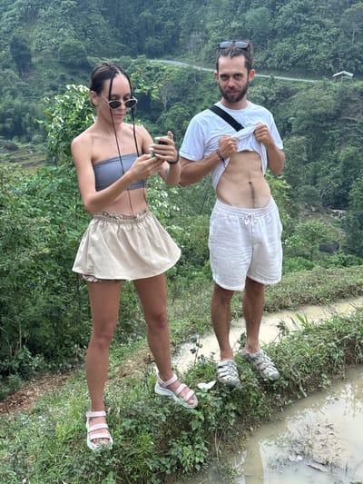 The image captures a man and a woman standing outdoors in a lush, mountainous landscape within Nậm Dịch Commune, Vietnam.
The man, on the right, has a beard, dark hair with some braids, and sunglasses pushed onto his head. He wears a white short-sleeved t-shirt, which he is pulling up with both hands to reveal his lean abdomen, showing a prominent vertical scar and muscle definition. He is also wearing white linen-style shorts and sturdy grey sandals. A black strap, possibly from a cross-body bag, runs across his chest. His feet are positioned near a muddy, water-filled channel bordered by dense green plants.
The woman, on the left, has dark braided hair and is wearing round sunglasses. She is dressed in a grey strapless crop top and a light beige, ruffled mini-skirt. She holds a smartphone with both hands, appearing to be interacting with it or taking a photo. Her legs are bare, and she wears light pink and white sandals, also standing near the edge of the vegetation.
The ground around them is a mix of dirt, green plants, and muddy water, suggesting a natural and possibly damp environment. In the background, rolling hills or mountains are covered in dense, vibrant green foliage, indicating a heavily forested area. A narrow road or path can be faintly seen winding through the distant slopes. The lighting appears soft and diffused, typical of an overcast day. No visible text is present in the image. The overall scene suggests the couple is exploring a scenic, rural area, possibly on a trek or outing.