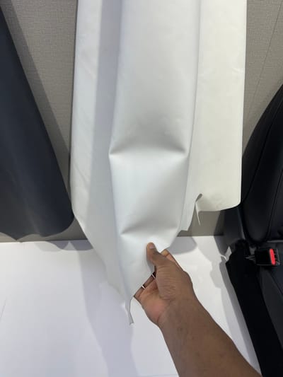 The image shows a person's hand holding a piece of white leather, which is positioned against a gray wall, a black leather sample, and a black car seat.  The white leather is smooth and appears to be a sample material. The person is examining the texture of the leather. The image was taken in a showroom in Hangzhou, China, at the location (30.272, 120.416).