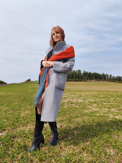The image depicts a woman standing in a field, likely located near Нечкино, Russia based on the presence of the Нечкинский национальный парк (Nechkinsky National Park). The woman has reddish-brown hair and is wearing a grey coat, black boots, and a pleated scarf with alternating orange and blue colors. She is positioned diagonally in the frame, facing the camera, with her hands clasped in front of her.
The field is covered with short green grass mixed with some dry patches of ground. In the background, there is a line of trees, suggesting a forest or wooded area. The sky is a light blue with scattered white clouds.