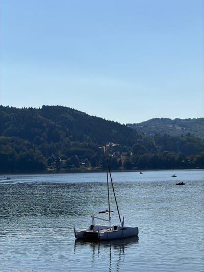 The image captures a serene scene on a body of water, likely the Lake Rożnowskie, near Gródek nad Dunajcem, Poland. A white catamaran sailboat is prominently featured in the foreground, anchored on the tranquil water. The vessel has two hulls and a central cabin structure, with a tall mast. In the background, a lush, tree-covered hillside rises steeply, dotted with houses nestled among the foliage. The sky above is a clear, bright blue, indicating fair weather. Several other smaller boats and watercraft, including what appear to be paddleboats and possibly other small recreational vessels, are visible on the lake in the middle distance, some with people aboard, suggesting a popular spot for water activities. The overall impression is one of a peaceful summer day in a scenic, natural setting.