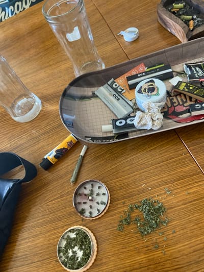 A wooden table contains drug paraphernalia. A rolling tray holds rolling papers, a container, and other items. Scattered marijuana sits next to a grinder containing more marijuana. A rolled joint and a lighter are also present. In the background, there is an ashtray with cigarette butts, along with two glass bottles.