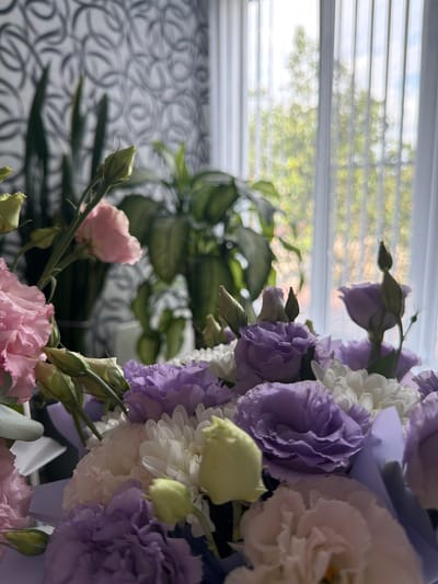 A close-up, slightly angled shot features a vibrant bouquet of flowers in the foreground. The bouquet consists of a variety of blooms including pink roses, purple lisianthus, and white chrysanthemums. Several green buds and leaves are also visible. The background is softly blurred, showing vertical blinds behind a window, with greenery and sunlight filtering through, suggesting an indoor setting with natural light. There are no people visible in the image. The location of Ust'-Dzhigutinka, Russia, is not discernible from the visual information presented in the image.