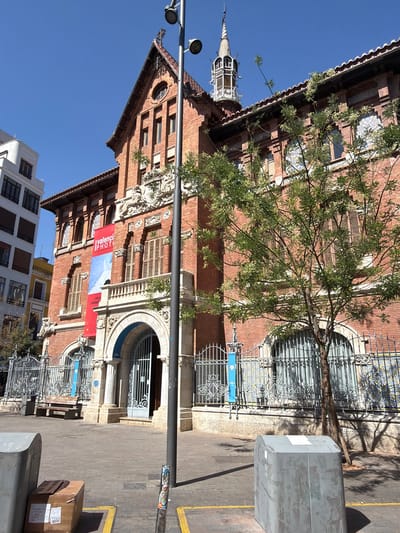 This is an eye-level, medium shot of the exterior of the Estació de França, a historic train station located in the city of Alboraia, Spain. The building is constructed of red brick with arched windows and ornate architectural details. A red banner with white text, reading "Valenciaport PHOTO" is hanging prominently on the facade. A mature tree with sparse green foliage stands in front of the building, partially obscuring the view of the windows and a decorative metal fence. The ground is paved with cobblestones, and there is a bench and trash receptacles visible in the foreground. The sky is clear and blue. There are no people visible in the image.