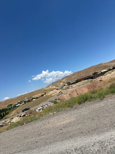 The image depicts a rural, mountainous landscape in Беш-Мойнок, Kyrgyzstan, under a clear blue sky with a few scattered clouds. The perspective is angled, with the sky dominating the left side and a dry, brown hillside on the right, suggesting a warm, possibly arid climate. A rough, unpaved road runs along the base of the hillside.
Along the roadside, there are scattered buildings, appearing to be houses, nestled into the slope. Some greenery, likely sparse vegetation or small trees, is visible around the buildings. A dark-colored car is parked by the side of the road, and a person in red clothing is standing near it. Power lines are strung across the scene, indicating some level of infrastructure. The overall impression is one of a remote, sun-drenched village in a mountainous region. The time of day appears to be daytime, with strong sunlight creating some shadows. There is no visible text in the image.