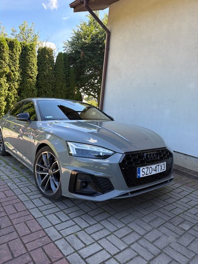 A grey Audi A5 coupe is parked on a herringbone-patterned brick driveway in front of a light-colored stucco house. The car's front is angled towards the viewer, with its license plate reading "SZO 4TX3". A row of tall green evergreen trees lines the left side of the image, and the sky above is blue with scattered white clouds. A gutter downspout is visible on the right side of the house. The scene appears to be a residential exterior setting.