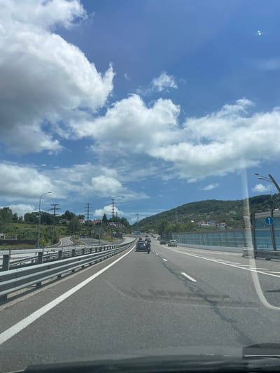 The image depicts a highway scene on a sunny day with fluffy white clouds in a blue sky. The perspective is from inside a vehicle, looking forward. The road is paved with asphalt and has clear white lane markings. A metal guardrail runs along the left side of the road, and a sound barrier made of blue glass is visible on the right. Several cars and a motorcycle are driving on the highway. In the background, there are green hills covered with trees and houses, suggesting a suburban or semi-rural area. Power lines and poles are also visible. The location is identified as Razdolnoye, Russia.