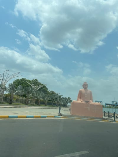 The image captures a full-body view of a large, light pink, seated Buddha statue situated outdoors. The statue is positioned on a rectangular pedestal of the same color and is surrounded by a paved area and a road. To the left of the statue, a line of palm trees and lush green trees are visible, along with decorative metal structures resembling palm fronds. In the background, several blue structures and fencing can be seen. The sky above is a clear blue with scattered white clouds. The scene appears to be a public space, possibly a park or monument in Ahmedabad, India, given the presence of the Buddha statue and the tropical vegetation.