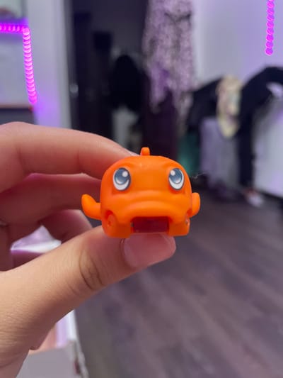 A person's hand is holding an orange toy with two blue eyes and a red mouth. The toy has small fins on its sides. The background is blurred, showing a room with purple lights. The image is taken indoors, possibly in a residential setting in Borisova Griva, Russia.
