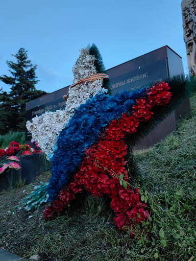A low-angle, medium shot captures a star-shaped wreath at the base of a monument in the Зуйское сельское поселение, Ukraine. The wreath is adorned with artificial flowers in the colors of the Russian flag: white, blue, and red, with a St. George's ribbon wrapped around the white section. The monument behind the wreath is dark and appears to be made of stone, with text inscribed on it that is partially visible and reads "2 ноября 1943 г. в Курск" and "жены жители:". A green coniferous tree is visible to the left of the monument, and other floral arrangements are scattered on the ground. The scene is set against a clear, pale blue sky, suggesting it is either early morning or late evening. The ground around the wreath is covered in grass and soil.