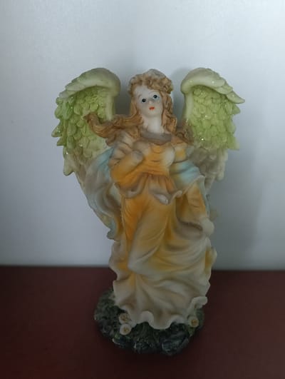 A vertically oriented, eye-level medium shot captures a figurine of a blonde-haired angel. The angel is depicted from the waist up, with its head tilted slightly upwards and to the right. It has blue eyes and wears a pale yellow gown with light blue accents. The angel's arms are crossed, and it has large, light green wings adorned with glitter. The figurine stands on a dark green base decorated with small white flowers. The background consists of a plain white wall. The figurine is placed on a dark red surface. There is no information about the location in Florești, Moldova.