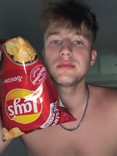 A close-up, waist-up, indoor, dimly lit selfie shows a young Caucasian man with light brown hair and blue eyes holding a bag of Lay's potato chips. He is shirtless and wearing a silver chain necklace. The bag of chips is red with a yellow and white Lay's logo, and appears to be a bacon flavor. Text on the bag is in Cyrillic script. The background is dark and appears to be a wall. The image is shot from a slightly high angle, looking down at the man.