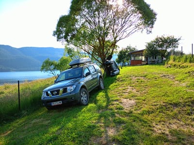A dark gray Nissan Pathfinder SUV, with a silver roof box, is parked at an angle on a grassy slope near a lake in Sarnitsa, Bulgaria. The car's license plate is PB 0756 MB. Behind the car, leaning against a large tree, is an inflatable raft. A small, rustic wooden house with a red trim and a screened porch is visible on the right side of the frame, further up the slope. The background features a wide expanse of water, likely a lake, and distant, tree-covered mountains. The sun is shining brightly, casting long shadows on the grass.