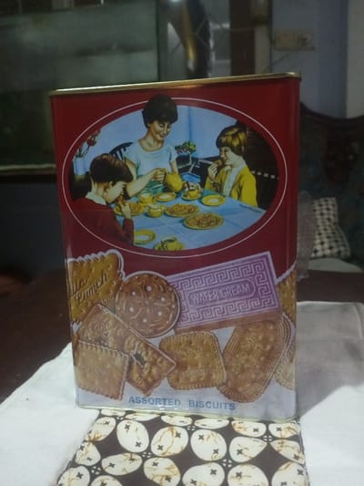 A metal rectangular biscuit tin, predominantly red with a gold rim, stands upright on a white fabric surface in a domestic setting within Buôn Ma Thuột, Vietnam. The tin's upper half features an oval illustration depicting an adult woman with dark hair, wearing a light-colored top, pouring from a yellow teapot into a cup while seated at a light blue-draped table. She is flanked by two children: a boy with dark hair to her left, holding a biscuit, and a girl with blonde hair and a yellow top to her right, eating a biscuit. The table holds multiple yellow teacups, saucers, a creamer, and plates of biscuits. The signature "Susan Pereygen" is visible on this illustration. The lower half of the tin displays images of various biscuit types, including "Villa Lunch Biscuit," a round patterned biscuit, a "Wafer Cream" biscuit with a Greek key pattern, and "K.G.B. Biscuit," with "ASSORTED BISCUITS" written at the bottom. The tin rests on a white cloth, which is placed over a dark brown surface featuring a white and brown patterned fabric underneath. The background is dimly lit and out of focus, revealing a large rectangular object with a greenish tint, possibly an aquarium, to the left, and dark wooden furniture, including a carved piece and a checkered fabric-draped item, to the right. A metallic, curved pipe-like object is visible in the upper-left background.