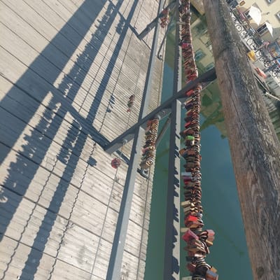 The image captures a close-up view of a wooden walkway adjacent to a body of water in Landshut, Germany. A prominent feature is a long chain adorned with numerous love locks, suspended from a metal railing. The wooden walkway is constructed from parallel planks and is partially covered by the shadows of an overhead structure. The water appears calm and reflects the surrounding environment. In the background, buildings and bicycles are visible, suggesting an urban setting. The overall scene conveys a sense of a public space, likely a bridge or a promenade.