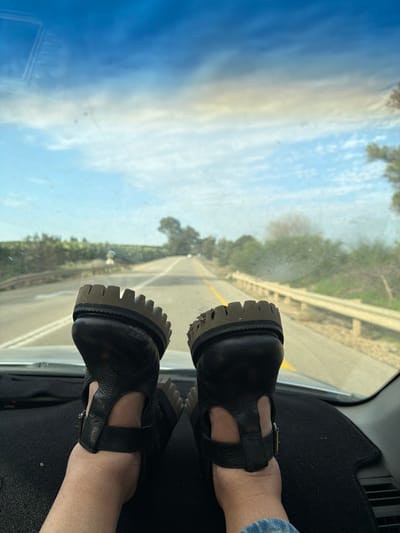 The image displays a first-person perspective from inside a vehicle, looking out over a road in the Hof Ashkelon Regional Council, Israel. Two feet are prominently propped on the black dashboard. These feet are clad in black, open-toed sandals with thick, serrated, dark-brown soles, one of which shows visible accumulated debris. The person's lower leg, partially visible on the right, wears light-wash denim. Through the vehicle's windshield, a two-lane asphalt road with a solid yellow center line extends into the distance, flanked by green vegetation and trees on both sides. A light-colored metal guardrail runs along the right side of the road. The sky overhead is partly cloudy with blue patches and scattered white clouds. A bridge-like structure is faintly discernible on the left side of the road in the mid-ground, and two small, distant vehicles are visible further down the road.