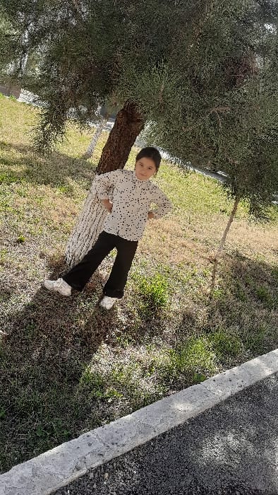 An image captured in Yangiariq, Uzbekistan, depicts a young female child standing outdoors during daylight. The child, dressed in a white long-sleeved shirt with black polka dots, black trousers, and white athletic shoes, leans with her back against a large tree trunk. Her hands are placed on her hips, and she faces forward with a slight smile. The tree features a textured, light-colored trunk and dense green foliage overhead. The ground consists of dry grass interspersed with patches of greener vegetation. To the right, a smaller tree with a thin trunk is visible. A concrete curb occupies the lower right foreground, separating the grassy area from a dark, granular surface, consistent with asphalt or gravel. Shadows are cast on the ground, indicating a bright light source.
