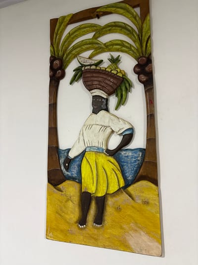 A vertical, painted wooden relief carving, located in Quimbaya, Colombia, is depicted mounted on a white wall. The artwork centrally features a dark-skinned female figure, dressed in a white top, yellow skirt, and white headwrap, standing barefoot. She balances a brown woven basket on her head, filled with various fruits including a pineapple, a watermelon slice, and multiple yellow spherical fruits. Her right hand rests on her hip. The figure is flanked by two brown palm trees with green fronds and coconut clusters. A textured blue section behind the figure represents water, while a yellow textured area below depicts sand. The carving displays a rustic, somewhat worn, painted finish.