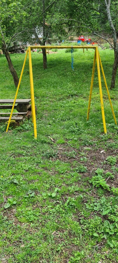The scene depicts an outdoor, rural or semi-rural setting in Ribaritsa, Bulgaria, characterized by overgrown green vegetation under an overcast sky. A bright yellow, metal swing set, consisting of a horizontal bar supported by two A-frame structures, stands in the midground; a single blue rope hangs from the center of the horizontal bar, but no swing seat is visible. To the left of the swing set, a rustic wooden structure resembling steps or a low bench, constructed from logs and planks and partially covered in moss, is situated. The ground is a mix of green grass, various weeds, scattered small white flowers, patches of bare earth, and numerous twigs and leaf litter. In the background, dense green deciduous trees dominate, partially obscuring a distant view. Through the foliage, a red tractor with prominent blue wheels is discernible on a patch of disturbed earth on the right. A light-colored building or structure is faintly visible through the trees on the left side of the background. No individuals are present in the image.