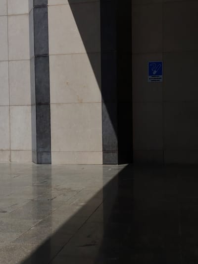 In Konigil, Uzbekistan, an outdoor scene depicts a section of a building's facade and the paved ground. A large rectangular column, composed of alternating bands of light and dark stone, extends vertically. The light-colored stone tiles are arranged in a grid pattern. To the right of the column, on a dark wall, a small blue and white sign is affixed. This sign features a stylized graphic of a cigarette and text that appears to be in a Cyrillic-based alphabet, accompanied by the English phrase "SMOKING AREA." A prominent diagonal shadow, cast by an unseen object, bisects the scene, falling across both the column and the reflective tiled ground. The ground itself is also tiled with light-colored stone, showing reflections of the architecture and the shadow. The overall impression is one of architectural detail under direct sunlight, creating stark contrasts between light and shadow.
