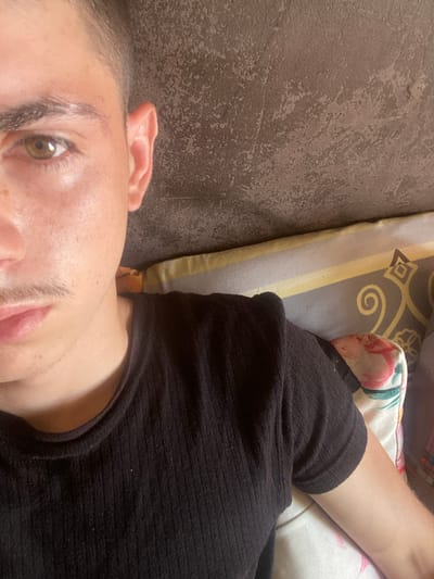 The image is a close-up selfie of a young male, taken indoors. The subject is positioned on the left side of the frame, with his head angled towards the camera. He has light brown eyes, visible eyebrows, and a faint mustache. His right ear is clearly visible. He is wearing a black, ribbed t-shirt.
The background consists of a textured wall on the upper portion of the image, and a pillow with a patterned cover on the lower portion. The pillow has yellow, gray, and possibly floral patterns. The lighting suggests it is daytime, with light illuminating the subject's face.
The location is identified as Gorna Malina, Bulgaria. However, the image itself does not contain any specific visual cues that would allow for a precise identification of this location within the city. The scene appears to be a domestic setting.