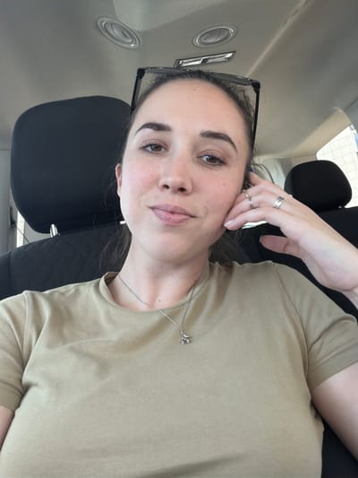 The image is a selfie taken by a young woman indoors, likely in a vehicle, given the presence of car seats and an air vent. She is looking towards the camera with a neutral expression, her right hand resting on her cheek. She is wearing a light brown t-shirt and a silver necklace with a small charm. She also has two silver rings on her left hand. Her dark hair is pulled back, and she is wearing black-rimmed glasses. The background is blurred, but shows hints of car interior and windows. The lighting suggests it is daytime. There is no visible text in the image.