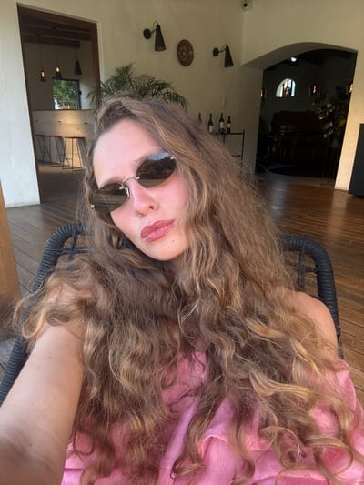 The image shows a person with long, wavy brown hair, wearing sunglasses and a pink top. They appear to be sitting in a chair on a wooden deck or patio. In the background, there's an interior space with a curved archway, tables, chairs, and wall decor, including sconces and possibly wine bottles. The scene is located in Stellenbosch Nu, South Africa. Through the archway, there are further interior details and a glimpse of the exterior, with light fixtures and an open window, suggesting a daytime setting. The person appears to be taking a selfie.