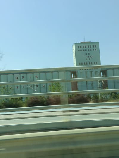 This media file captures a view of an industrial facility in Konya, Turkey. The primary subjects are a tall, light green tower building and a series of silo-like structures. The tower building has numerous rectangular windows arranged in rows and a prominent "TMO" logo in dark red letters. The silos are lighter in color and have "OFİS" and "DOSTİDUR" written in dark red letters across them, suggesting they are part of an office and storage facility.
The scene appears to be outdoors on a clear, sunny day, indicated by the bright blue sky and the strong shadows cast by the structures. The perspective suggests the photo was taken from a moving vehicle, likely a car or bus, as evidenced by the blurred foreground and the presence of horizontal bars, resembling a guardrail or window frame, across the image. The motion blur adds a dynamic element to the otherwise static industrial landscape.
The overall mood is neutral, focusing on the architectural and textual elements of the industrial site. The location is specifically identified as Konya, Turkey, which provides geographical context for these structures. The clear weather and daytime setting are consistent with an active industrial zone.