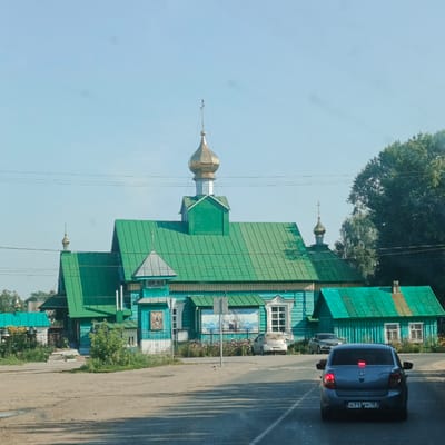 The image shows a bright green wooden building, likely a church, in Kambarka, Russia. The building features ornate white trim below the roofline and has multiple gabled sections covered in green metal roofing. A golden dome with a cross sits atop a central tower, and a smaller dome with a cross is visible on another section of the roof. The church is situated on the side of a dirt and asphalt road. In the foreground, a gray sedan with its brake lights illuminated is stopped on the asphalt. Behind the church, there are trees and a clear blue sky. Several cars are parked near the building, and some vegetation, including flowers and bushes, is visible along the front of the church. The overall scene suggests a sunny day in a rural or small-town setting.