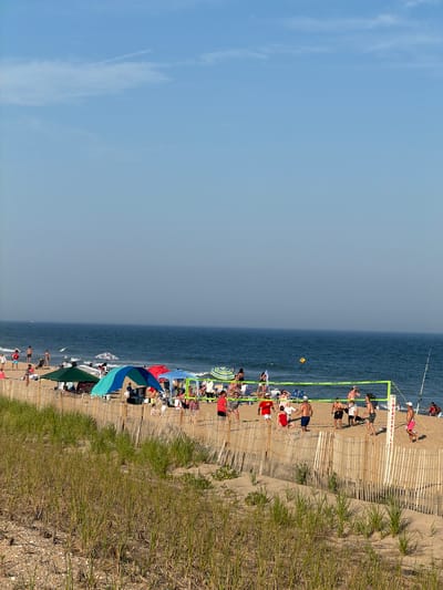 In Montauk, United States of America, a sunny day unfolds on a crowded beach. The ocean, a deep blue, stretches to the horizon under a clear sky with a few wispy clouds. The sandy shore is dotted with numerous beachgoers enjoying various activities. A group of people is engaged in a lively game of beach volleyball, with players positioned on either side of a bright green net. Nearby, beach umbrellas and tents provide shade for sunbathers. A wooden fence runs along the edge of the beach, separating it from a sandy dune covered in tall, dry grass. The scene captures a typical summer day at the beach, full of recreation and relaxation.