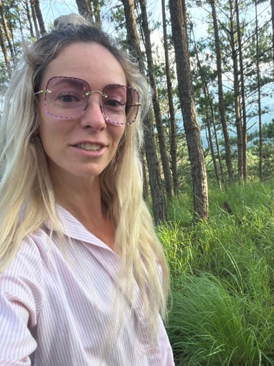A woman with blonde hair, wearing oversized pink-tinted square glasses and a pink and white striped button-up shirt, stands in a forest with tall pine trees and lush green grass. The background shows more trees and a hint of the sky. The woman is looking towards the camera and smiling. The location appears to be Xuan Truong - Da Lat, Vietnam.