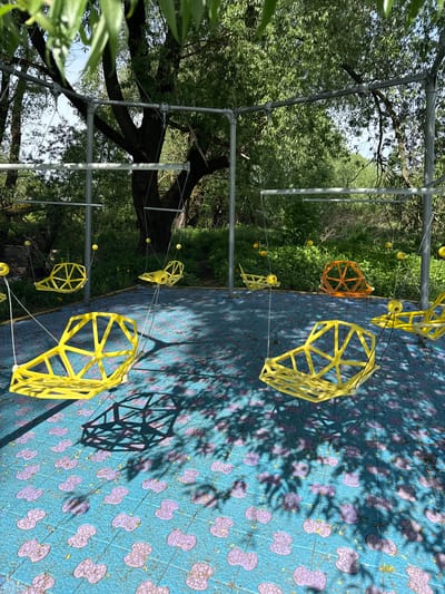 A partially inverted eye-level outdoor shot taken in Синьково, Russia, presents a geometrically themed playground or park installation against a natural backdrop of trees and vegetation. The image is rotated counterclockwise, so the intended “ground” appears on the right.
In the foreground and central area, several geometric structures made of yellow and orange materials are suspended or placed on a blue surface patterned with purple clover-like shapes. These structures resemble hollow polyhedrons or partial geodesic domes, some casting stark shadows on the patterned ground. They are connected with thin, white strings to silver metal poles. The geometric shapes appear to be part of a playground installation.
In the background, a green landscape is visible, including trees, plants, and hints of other park elements. Metal poles support the structures. The light suggests it is daytime with clear skies.