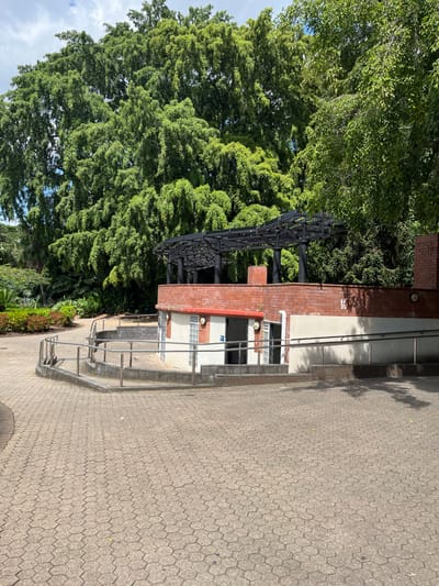 The image displays an outdoor scene featuring a public building, a large tree, and a distinctive black metal structure under bright, clear daylight.

The building, likely an amenities block, is constructed with red brick on its lower section and a lighter, possibly rendered, material on its upper half. It has multiple entrances, one of which is accessed via a ramp equipped with silver metal handrails. Several small, circular outdoor lights are affixed to its facade, and a white drainage pipe runs vertically down one wall. A rectangular window section features glass blocks, and the text "7B" is visible in dark numbers on the lighter-colored wall.

To the left of the building, a very large tree with a dense, verdant green canopy dominates the frame, suggesting a mature park or garden setting. Smaller shrubs and plants form a garden bed at the base of this tree and along the front of the building.

Above the building and partially extending into the tree's foliage, an intricate black metal structure is prominent. It features an angular, grid-like design and appears to be a sculpture or a unique architectural element.

The ground area in front of the building and stretching into the foreground is paved with grey hexagonal interlocking pavers. Shadows are cast on this surface, indicating a sunny day. Patches of blue sky with some white clouds are visible through the tree canopy in the upper left. The overall scene suggests a well-maintained public space, consistent with an urban park or facility in Brisbane City, Australia. No people are visible, and no specific event is taking place.
