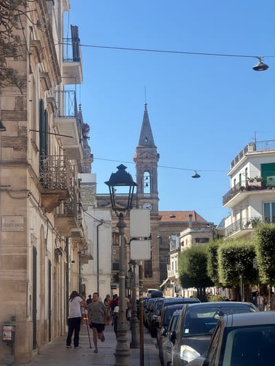 This image depicts a street scene in Alberobello, Italy, under a clear blue sky. The left side of the frame features a building with a light-colored facade and ornate balconies. A street sign reading "VICO SOLDATO IGNTO" is visible. On the right side, a white building with balconies and potted plants is situated. A prominent bell tower with a clock face rises in the background, partially obscured by other buildings and trees. The street is lined with parked cars. In the foreground, several people are walking along the sidewalk. One person in a white t-shirt and dark pants walks away from the camera, while another in a grey t-shirt and shorts runs towards the camera, holding a red pole. Other individuals are visible in the background. A street lamp with an ornate design stands prominently in the middle ground. The overall impression is that of a sunny day in a historic Italian town.