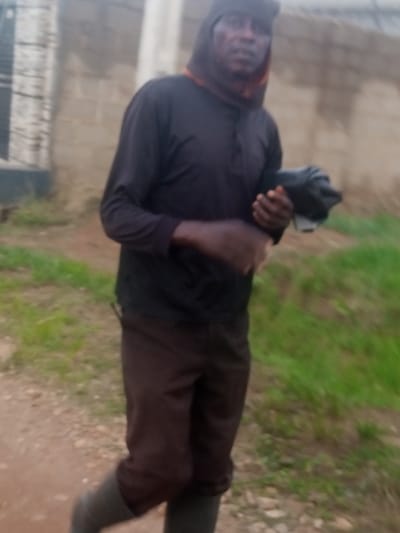 A person, likely male, is walking outdoors on a dirt path. He is wearing a dark hooded sweatshirt with an orange trim visible at the neck, dark pants, and grey boots. The person is holding a dark object in his right hand, which is partially obscured. The background consists of a light-colored brick wall on the left and grassy areas on the right. The lighting suggests it might be daytime, possibly overcast or late afternoon given the somewhat muted tones. The setting appears to be rural or semi-urban, consistent with a location like Russo, Nigeria. There is no visible text in the image.