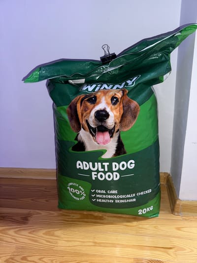 This is a media file of a large bag of dog food. The bag is primarily green with white text and an image of a dog's face. The text on the bag includes "WINNY", "ADULT DOG FOOD", "COMPLETE AND 100% BALANCED FOOD", "ORAL CARE", "MICROBIOLOGICALLY CHECKED", "HEALTHY SKIN&HAIR", and "20KG". The dog's face appears to be a beagle or similar breed, with its tongue sticking out. The bag is partially open and folded over, held by a black binder clip. The background consists of a wooden wall on the left and a white wall on the right. The lighting suggests an indoor setting, possibly a room with natural light from a window. The image is rotated, with the bag appearing to be upright in the scene. The setting appears to be domestic, likely a home. There are no people or animals other than the image on the bag.
