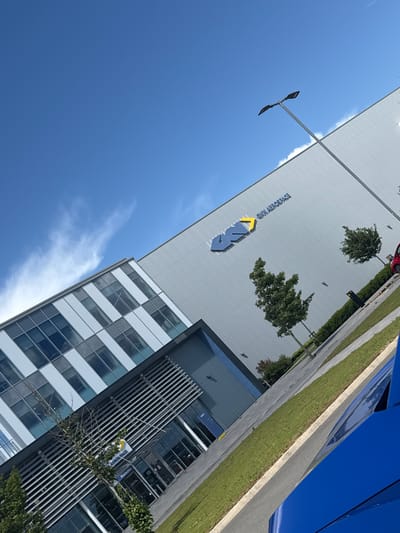 The image shows the GKN Aerospace facility in Filton, United Kingdom, on a clear day with a blue sky and scattered clouds. The building's facade is a mixture of corrugated metal and glass windows. The GKN Aerospace logo is prominently displayed on the upper section of the building. In the foreground, a bright blue vehicle is partially visible. The scene includes neatly manicured green lawns, trees, and paving stones. The perspective is from a low angle, looking upwards at the building and sky.