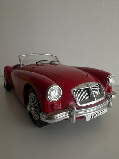 A red convertible toy car is positioned on a light-colored surface, with a plain, light gray wall in the background. The car is facing towards the viewer, slightly angled to the right. It has a chrome grille, headlights, and bumper. A license plate on the front bumper reads "UMG 400". The car's spoked wheels are visible. The lighting is even, casting soft shadows. There are no people or other objects in the image. This scene is not in Lapės, Lithuania.
