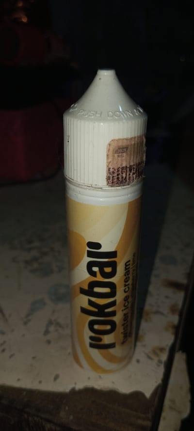 A bottle of I-okbar "twister ice cream" flavor e-liquid stands upright. The bottle has a white childproof cap that says "PUSH DOWN & TURN." A label with the date "7/2025" and the acronym "REL" is partially peeled. The yellow and white swirl design wraps around the bottle's body. A dark and out-of-focus background obscures the surroundings. The surface underneath the bottle shows dark stains and markings.