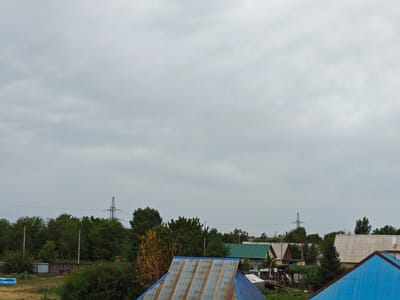 An overcast, grey sky fills the upper portion of the image, with a line of trees separating the sky from a residential area below. Several houses with different colored roofs, including green and corrugated metal, are visible amongst the trees. Electrical pylons can be seen in the distance. In the foreground, a bright blue tarp is draped over a structure, and a rusty corrugated metal roof is angled upwards. A person in a green shirt and dark pants is walking across a grassy area towards the left of the frame, near a blue inflatable pool. The overall scene depicts a rural or semi-rural residential area under a cloudy sky.
