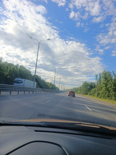 This media file captures a scenic view from a car driving on a highway in Бавыкино, Russia. The scene is dominated by a clear blue sky with scattered white clouds, suggesting fair weather. The time of day appears to be morning or late afternoon, indicated by the soft lighting and the long shadows cast on the road.
The highway itself is a prominent feature, with multiple lanes and clear lane markings. On the left side of the road, a metal guardrail separates the highway from a wooded area. A large white truck is visible in the distance on the left, suggesting ongoing traffic. On the right side of the highway, a dense forest with lush green trees lines the road.
A red car is driving ahead of the viewpoint car, indicating movement and travel. The dashboard of the car from which the image is taken is partially visible in the foreground, showing a brown-colored upper section and a dark gray lower section with a curved black line, possibly a wire or a part of the car's interior.
Several tall lampposts are spaced along the highway, characteristic of road infrastructure. The overall atmosphere is one of peaceful travel through a natural landscape. There is no visible text in the image.