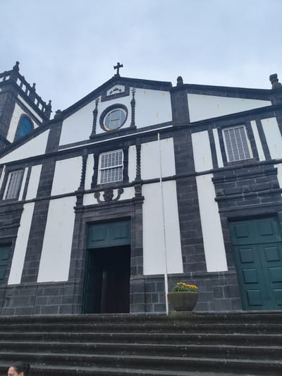 The image shows the front facade of a church in Ribeira Grande (Conceição), Portugal. The church has a striking black and white striped design on its upper facade, with dark grey stone on the lower facade and around the windows and doors. A tall, narrow tower with a cross on top is visible to the left. Several windows are visible, including a circular one above the central entrance. The main entrance is a large, arched doorway with dark green double doors, one of which is open. To the right of the entrance, another set of dark green double doors is visible. A stone staircase leads up to the entrance. In front of the entrance, on the staircase, is a stone planter with yellow flowers. The sky above is overcast. A woman is partially visible in the lower left corner of the image, standing on the staircase.