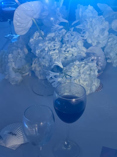 This is a close-up, overhead shot of a table setting with a bouquet of white flowers, likely hydrangeas and an anthurium. The bouquet is arranged in shells and pearls. Two wine glasses are prominent; one is empty and the other contains red wine. A seashell with the inscription "Maps" is placed next to the empty glass. The overall lighting is a cool blue, creating a serene and possibly evening ambiance. The image depicts a still life arrangement on a table, likely part of a decorative display or a prepared table for an event. There is no indication of the city of Osipovo, Russia, in the image.