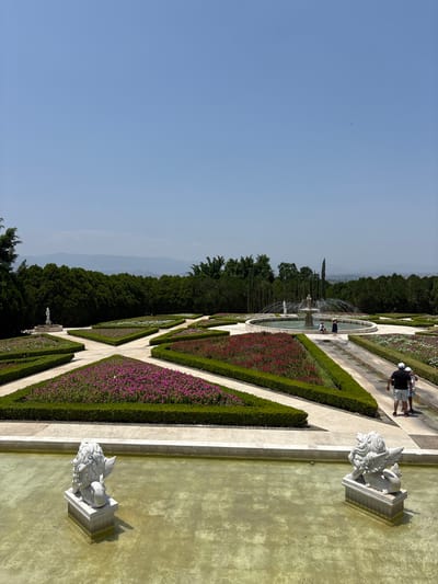 A slightly rotated, eye-level outdoor shot depicts a formal garden with geometric flowerbeds and pathways, likely situated in Tehuixtla, Mexico, given the prompt. A group of people is walking along one of the pathways in the background. Two stone statues, possibly depicting lions, are positioned along the edge of a rectangular pool of water. The water is tinted green. The garden is bordered by a dense line of trees. The sky is clear and blue.