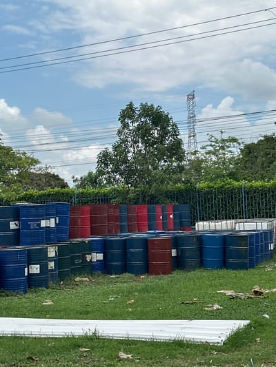 The image shows an outdoor scene with stacks of metal barrels in Juan Mina, Colombia. The barrels are predominantly blue, with some red and green ones interspersed. They are arranged in rows, with some stacked two high. In the background, there is a fence, lush green trees, and a transmission tower against a partly cloudy sky. The ground is covered with grass and some scattered debris. There are no people or animals visible in the image. The overall impression is of an industrial or storage area.