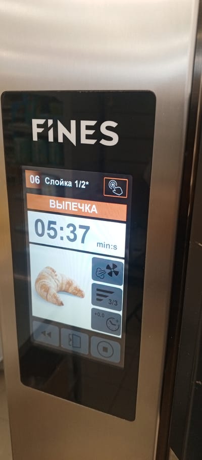 This is a close-up, eye-level, medium shot of a digital display on a stainless steel appliance. The appliance has the brand name "FINES" prominently displayed in white, uppercase letters on a black background at the top. The display itself is a touchscreen interface.

The screen shows a program selection labeled "06 Слойка 1/2*", which translates to "06 Puff pastry 1/2*". Below this, in larger, bold text, is "ВЫПЕЧКА", meaning "BAKING". The timer is set to "05:37", indicating 5 minutes and 37 seconds remaining in the baking cycle.

To the left of the timer is an image of a croissant. To the right of the croissant image are three icons. The top icon depicts a fan with airflow, suggesting convection. The middle icon shows three horizontal lines, possibly indicating the stage of baking or a timer segment. The bottom icon is a clock with a plus sign, indicating some form of time adjustment or a timer function.

Below the main display area are control buttons. On the bottom left, there are two arrow icons pointing left, likely for navigation or rewind. In the center is an icon that resembles a folder or a document, possibly for saving or loading programs. On the bottom right, there is a circular button with a square inside, which is likely a stop or pause button.

The appliance appears to be a commercial oven or similar kitchen equipment. The image does not contain any information that would allow for its location to be identified within Zhirnovsk, Russia, or any other city. There are no people or interactions visible in the image. The environment appears to be indoors, likely in a commercial kitchen or food service setting, given the nature of the appliance. The overall lighting is bright, highlighting the metallic surface of the appliance and the illuminated screen.